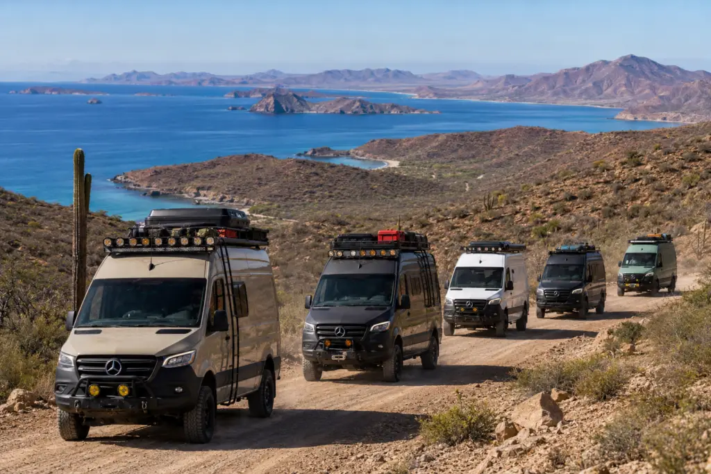 Five custom Mercedes-Benz Sprinter vans, in tan, black, white, and green, drive on a dirt road. A saguaro cactus is on the left. In the background, a vast blue ocean with rocky islands and arid mountains under a clear sky.