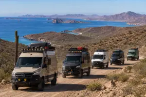 Five custom Mercedes-Benz Sprinter vans, in tan, black, white, and green, drive on a dirt road. A saguaro cactus is on the left. In the background, a vast blue ocean with rocky islands and arid mountains under a clear sky.