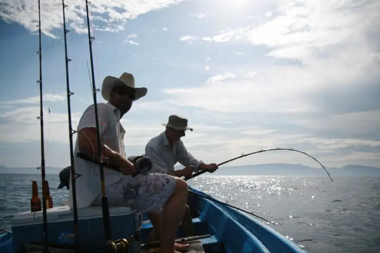 Two men fishing from a blue boat on sparkling water under a bright sky, one wearing a cowboy hat and sunglasses, the other a bucket hat and striped shirt.