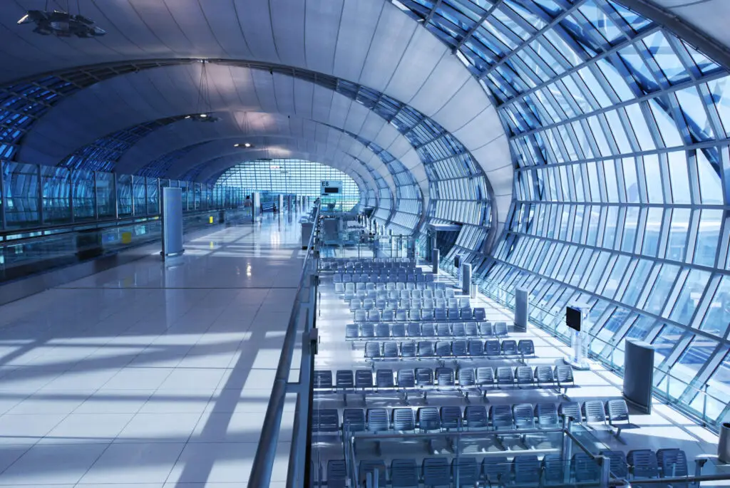 A wide, high-angle shot of an empty, modern airport terminal with an arched ceiling, extensive glass walls, a long moving walkway, and rows of empty seating.