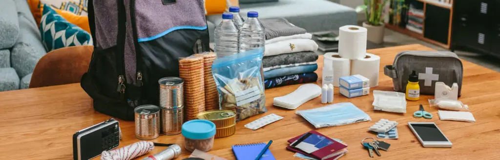 An emergency preparedness kit laid out on a wooden table, including a backpack, water, food, first aid supplies, hygiene products, clothes, and documents.