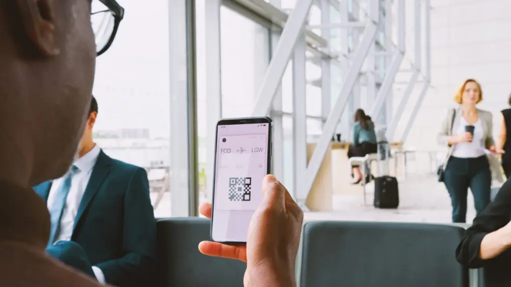 A person holds a smartphone displaying a digital boarding pass with a QR code and flight details in an airport lounge.