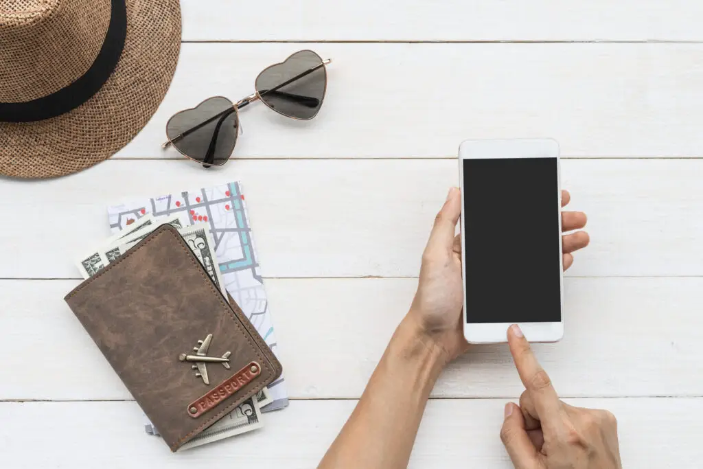 Overhead view of travel items on a white wooden table: a straw hat, heart-shaped sunglasses, a passport holder with money and a map, and hands holding a smartphone.