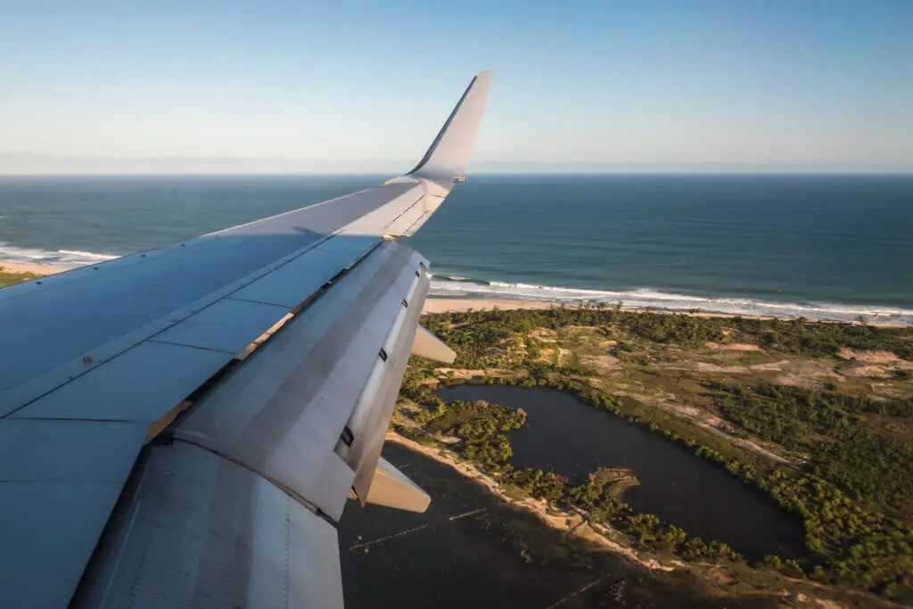 New Flights & Airport Expansion 1 Aerial view from an airplane window showing a wing, a sandy beach with waves, green vegetation, a dark lagoon, and the ocean under a clear sky.