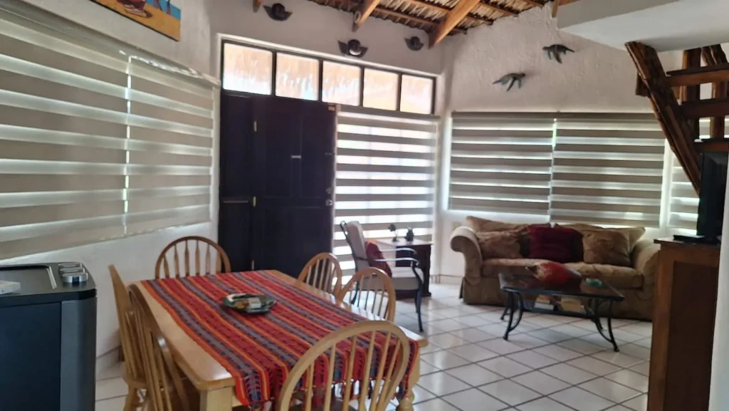 El Palmar Casa — Club Rental in Loreto 4 An interior shot of a living and dining room with a wooden table, striped tablecloth, brown sofa, and zebra blinds on the windows. A thatched ceiling and tiled floor are visible.