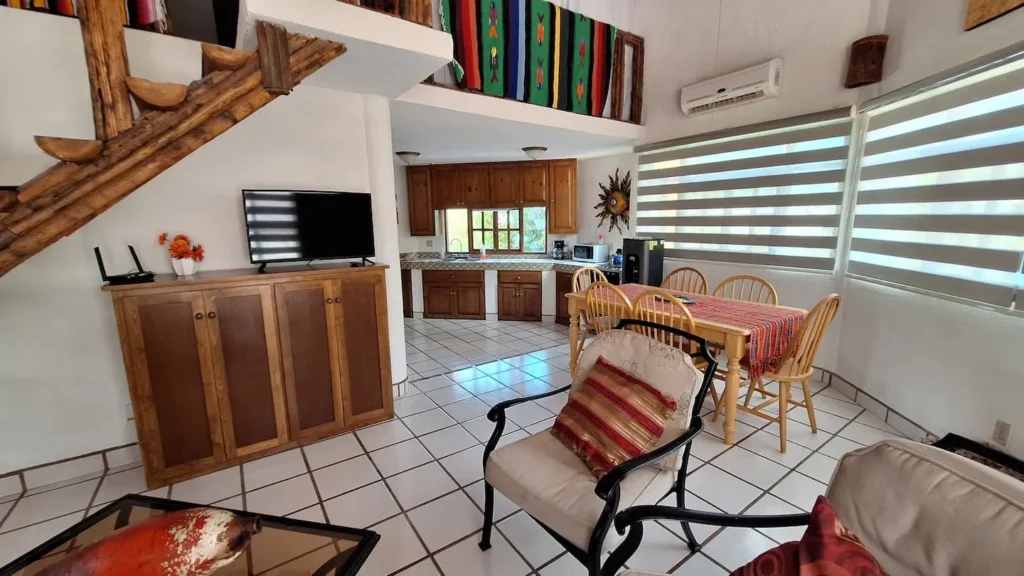 El Palmar Casa — Club Rental in Loreto 6 An open-concept living room, dining area, and kitchen with white tiled floors, rustic wooden staircase, and colorful accents.