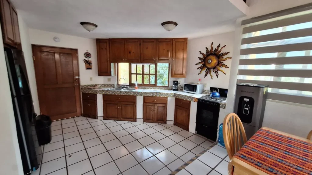 El Palmar Casa — Club Rental in Loreto 3 A kitchen with wooden cabinets, tiled countertops, a double sink, a black stove, and a large sun decoration on the wall. A refrigerator, water dispenser, and dining table are also visible.
