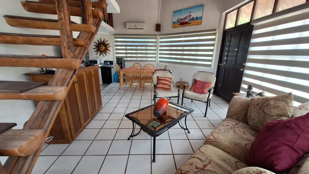 El Palmar Casa — Club Rental in Loreto 5 An open-concept living and dining room with a rustic wooden staircase, white tiled floor, patterned sofa, armchairs, glass coffee table, and a wooden dining set. Windows with horizontal blinds and a dark door are visible in the background.