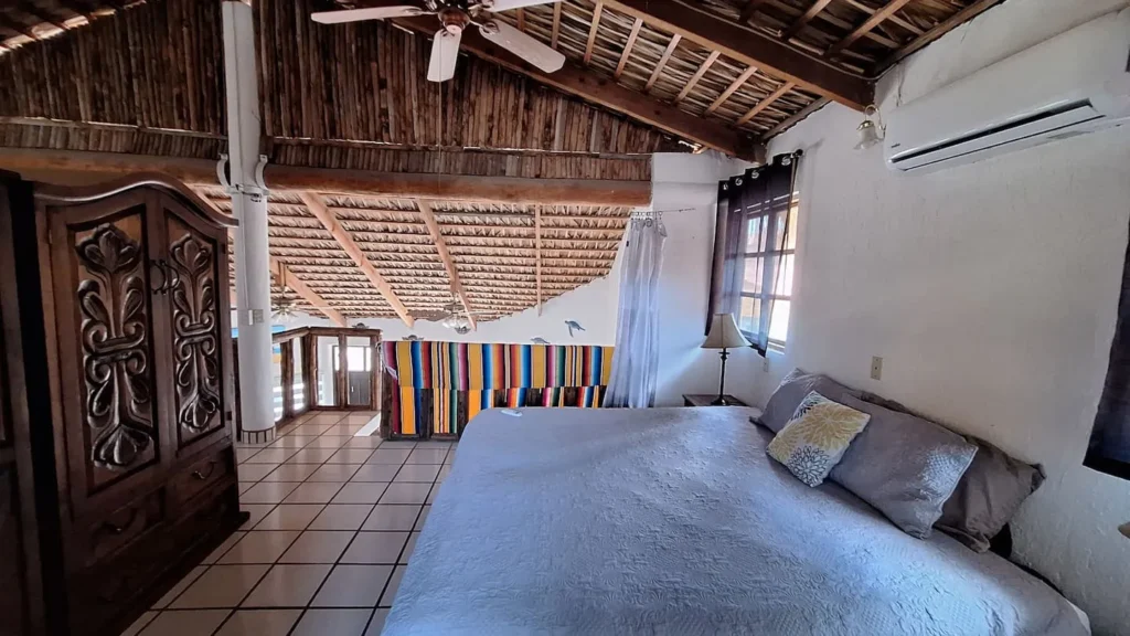 El Palmar Casa — Club Rental in Loreto 7 A rustic bedroom with a large bed, carved wooden armoire, thatched ceiling, and a colorful striped blanket as a headboard.