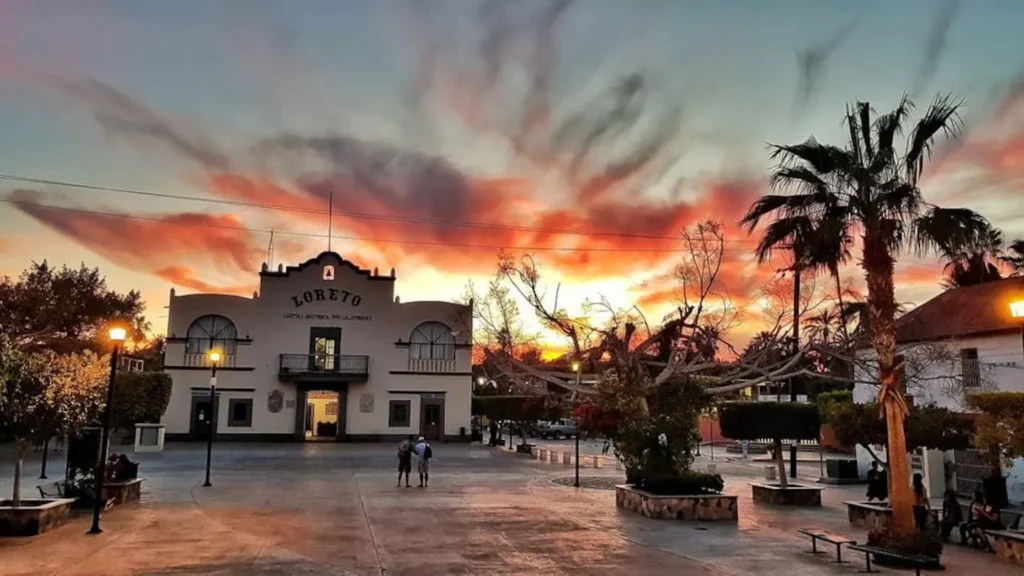 A town plaza at sunset, featuring a historic building with 'LORETO' written on it, a tall palm tree, and illuminated lampposts under a sky filled with dramatic orange and red clouds.