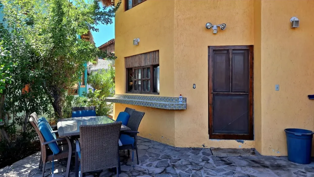 El Palmar Casa — Club Rental in Loreto 2 Outdoor patio with a glass-top dining table and wicker chairs with blue cushions, next to an ochre building with a window, screen door, and a tiled counter.