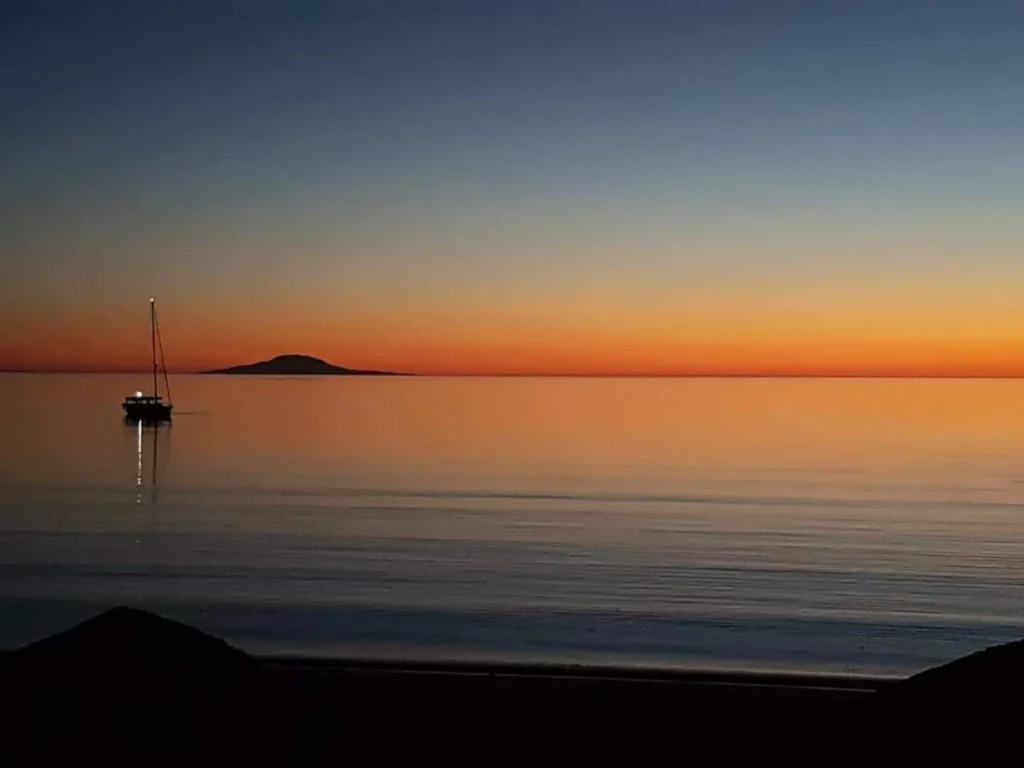 A sailboat on calm water at sunset, with a distant island silhouetted against an orange and blue sky.