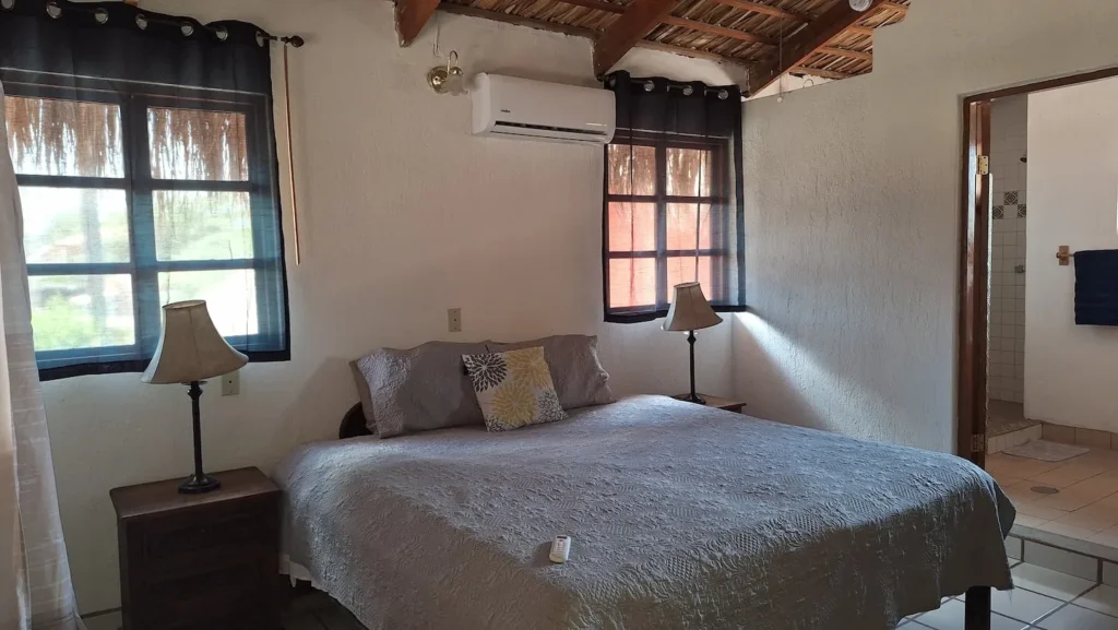El Palmar Casa — Club Rental in Loreto 8 A rustic bedroom with a large bed, two windows with sheer curtains, and an open doorway to a tiled bathroom.